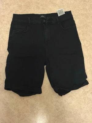 Svarta shorts från ONLY - Svarta shorts från ONLY med klassisk femficksdesign och uppvikta benslut. De har normal passform och midja, samt bälteshällor och knappstängning. Perfekta för sommaren och enkla att matcha med olika stilar.