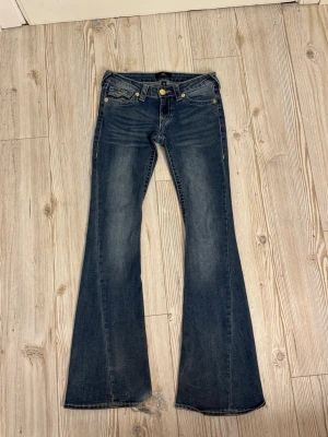 Blå bootcut jeans från True Religion i storlek w24  - Säljer ett par blå bootcut jeans från True Religion med klassiska kontrastsömmar, låg midja, guldknappar på bakfickorna och gula detaljer. Jeansen är i bra skick förutom lite slitningar vid fötterna!🤍