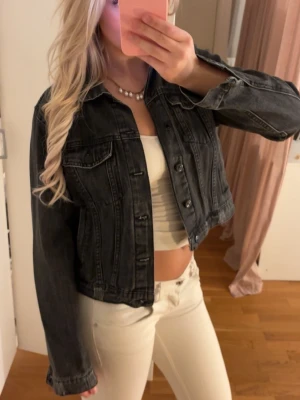 Svart jeansjacka med klassisk look - Säljer en svart jeansjacka med klassisk passform och knappar framtill. Jackan har två bröstfickor med lock och knapp samt en croppad längd som ger en trendig vibe. Perfekt att slänga över en topp för en cool och avslappnad stil.