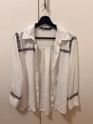 Vit skjorta med broderade detaljer Zara - Vit skjorta från Zara med färgglada broderade band längs axlar och ärmslut. Skjortan har klassisk krage och långa ärmar. Materialet känns lätt och luftigt, perfekt och avslappnad look.