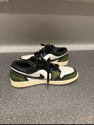 Nike Air Jordan 1 Low svart/vit/grön - Snygga Nike Air Jordan 1 Low sneakers i svart, vitt och grönt med klassisk swoosh på sidan. Skorna har lågt skaft, svarta skosnören och en vit sula. Ovandelen är i läder och mesh för en fräsch och sportig look.