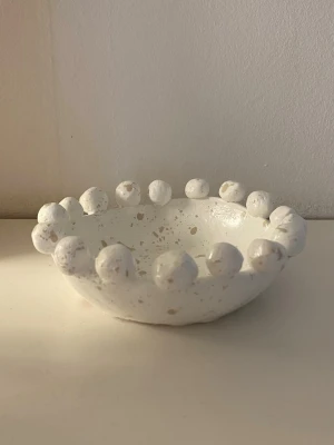 Vit skål med bollar - Handgjord skål i lera med runda bollar längs kanten och små beige prickar över hela ytan. Skålen har en rund form och passar perfekt som dekoration eller förvaring av småprylar.