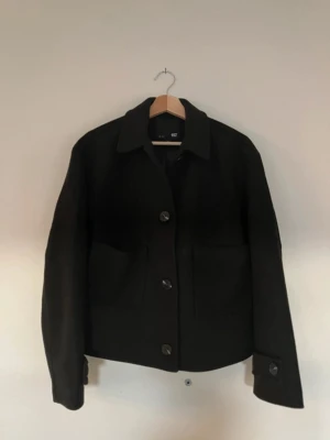 Svart jacka/kappa - Stilren svart cardiganjacka från Lager157 med klassisk krage och stora knappar framtill. Jackan har två rymliga fickor och raka ärmar med knappdetalj vid ärmslut. Perfekt för lager på lager och enkel att matcha med olika outfits.