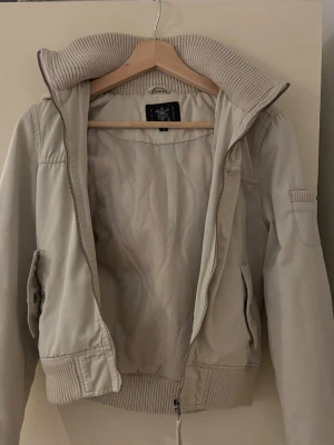 Beige bomberjacka med ribbad krage - Snygg beige bomberjacka från Eve of Eden med bred ribbad krage och muddar. Jackan har dragkedja framtill, två fickor med lock och en extra ficka på ärmen. Insidan är quiltad för extra komfort och värme. Perfekt för dig som vill ha en stilren och trendig look.