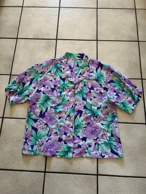 Lila blommig skjorta med knappar storlek L, Bystomkrets 106 cm - Säljer en färgstark skjorta med blommigt mönster i lila, grönt och vitt. Skjortan har klassisk krage, korta ärmar och knappar framtill. Perfekt för dig som vill sticka ut med en cool och somrig look.