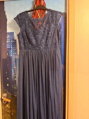 Marin Blå balklänning med spetsdetaljer - Elegant marin blå balklänning med vackert spetsliv och blommigt mönster. Klänningen har knappar i ryggen, rundad halsringning och korta ärmar. Kjoldelen är i mjukt, fallande tyg som ger en romantisk känsla. Perfekt för speciella tillfällen. Stl XL