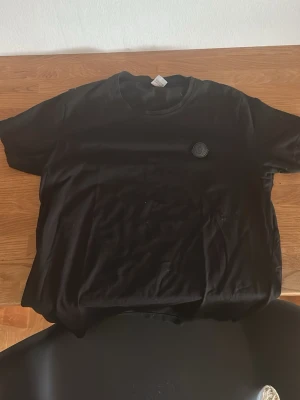 Svart Moncler t-shirt i bomull - Snygg svart t-shirt från Moncler med rund hals och diskret logga på bröstet. Tillverkad i mjuk bomull och har en klassisk passform med korta ärmar. Perfekt till jeans eller shorts för en clean och stilren look.