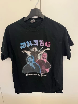 Svart Drake t-shirt Champagne Papi - Cool svart t-shirt med tryck av artisten Drake och texten 'Champagne Papi'. T-shirten har rund hals och korta ärmar. Perfekt för dig som gillar musik och streetwear. I bomull