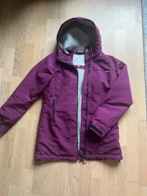 Vinröd vindjacka från Sail Racing - Snygg vinröd vindjacka från Sail Racing med huva och svart dragkedja. Jackan har meshfoder, justerbara muddar med kardborre och en praktisk ficka med dragkedja på sidan. Perfekt för blåsiga dagar och riktigt skön passform. Frakten kan variera i pris.