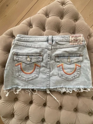 Ljus jeanskjol från True Religion - Säljer en ljusblå jeanskjol från True Religion med rå kant och slitna detaljer. Kjolen har två bakfickor med färgglada broderier och knappstängning. Klassisk True Religion-lapp bak i midjan. 