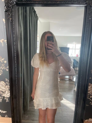 Vit kort klänning med puffärm - Söt vit kortklänning med puffärmar, broderade blommor och volangkant. Klänningen har v-ringning, öppen rygg med knyt och är tillverkad i ett lätt tyg som passar perfekt till sommaren.