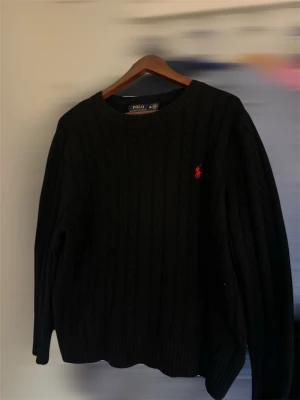 Svart kabelstickad tröja Polo Ralph Lauren - Kabelstickad svart tröja från Polo Ralph Lauren med rund halsringning och den klassiska röda logotypen broderad på bröstet. Tröjan har ribbade muddar vid ärmslut och nederkant. Perfekt för dig som gillar stilrena och tidlösa plagg.