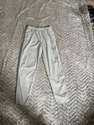 Nike ljusgrå mjukisbyxor med logga - Säljer ett par ljusgrå mjukisbyxor från Nike med elastisk midja och diskret Nike-logga på benet. Byxorna har raka ben och en grå rand längs sidorna. Perfekta för chill dagar eller träning. Materialet känns lätt och bekvämt.