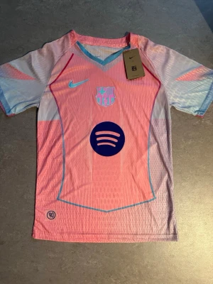 Barça x Nike rosa matchtröja - Snygg FC Barcelona matchtröja från Nike i rosa och ljusblått med klubbmärke, Nike-logga och Spotify-logga på bröstet. Tröjan har korta ärmar, V-ringning och är tillverkad i lätt, ventilerande funktionsmaterial. Perfekt för fotbollsträning eller support.