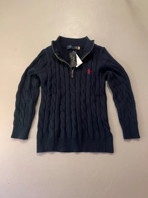 Mörkblå kabelstickad tröja Polo Ralph Lauren - Mörkblå kabelstickad tröja från Polo Ralph Lauren med halv dragkedja och ribbade muddar. Tröjan har det klassiska röda Polo-logot broderat på bröstet och är tillverkad i mjukt material. Perfekt för en stilren och avslappnad look.
