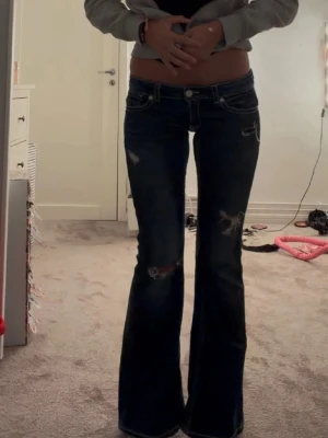 Mörkblå bootcut jeans med slitningar - Säljer ett par mörkblå bootcut jeans med slitna detaljer och låg midja. Jeansen har vita kontrastsömmar och klassisk femficksdesign. Perfekta för en trendig och avslappnad look.