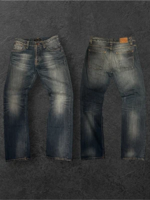 Vintage Nudie Jeans - Säljer ett par klassiska blå jeans från Nudie Jeans med snyggt slitna detaljer och raka ben. Jeansen har fem fickor, orange sömmar och en läderpatch med logga bak i midjan. Tillverkade i slitstark denim som ger en cool vintagekänsla. HELT NYA!