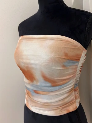 Batikmönstrad bandeau tubtopp - Trendig bandeau tubtopp med batikinspirerat mönster i ljusa toner av beige, orange och lite blått. Toppen är axelbandslös och har en tight passform med lätt rynkade sidor. Perfekt för sommarens alla outfits.