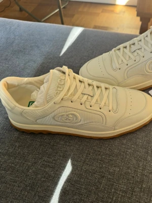 Gucci vita sneakers med beige sula - Säljer ett par stilrena vita sneakers från Gucci med beige gummisula. Skorna har klassisk låg profil, rundad tå och detaljer i både läder och textil. Gucci-loggan är tydligt broderad på sidan. Perfekta för dig som vill ha en clean och exklusiv look.