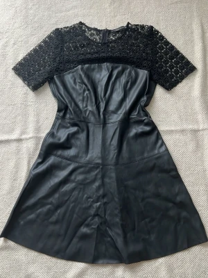 Svart klänning med spets och skinn - Snygg svart klänning med överdel i blommig spets och nederdel i skinnimitation. Klänningen har korta ärmar, rund halsringning och dragkedja i ryggen. Perfekt för dig som vill ha en edgy men feminin look.