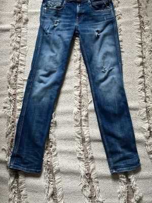 Blå slitna jeans från Replay - Säljer ett par blå jeans från Replay med slitningar och hål på framsidan för en trendig look. Klassisk femficksmodell med raka ben och snygga detaljer på bakfickorna. Jeansen har en läderpatch med Replay-logga bak i midjan. Perfekta för dig som gillar en avslappnad stil.
