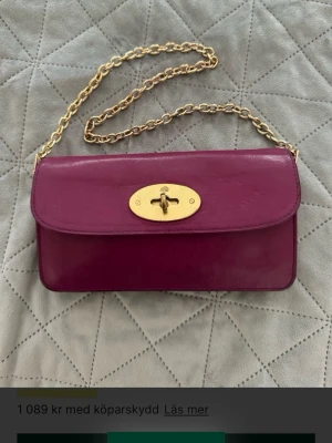 Rosa ( mörkrosa)  handväska/Pochette med guldkedja från Mulberry - Snygg lila handväska/pochette i glansigt läder med guldfärgad kedjerem och vridlås framtill. Väskan har en rektangulär form och elegant design, perfekt för dig som vill sticka ut. Guldfärgade metalldetaljer ger en lyxig känsla. Finns både en längre samt en kortare kedja. Hårvatten används som crossbody och en kortare version