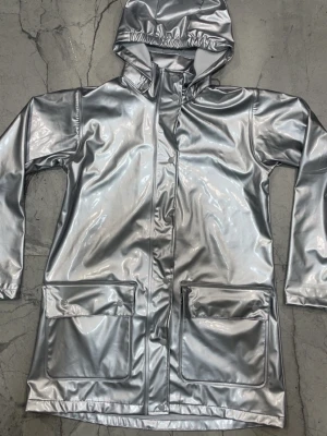 Silverfärgad regnjacka med huva - Cool silverfärgad regnjacka med huva och stora fickor framtill. Jackan har en blank finish och är tillverkad i ett vattenavvisande material som håller dig torr. Perfekt för att sticka ut i regnet och hålla stilen. Stängs med dragkedja och tryckknappar.
