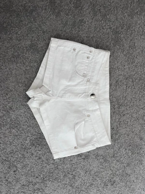 Vita lågmidjade jeansshorts - Säljer ett par vita lågmidjade jeansshorts med klassisk femficksdesign och knappgylf. Shortsen har en liten patch med logga bak och är perfekta för sommaren. 