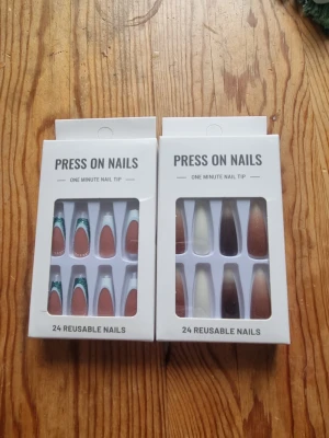 Press-on naglar i brunt och vitt - Två förpackningar press-on naglar med 24 återanvändbara naglar i varje. Ena setet har bruna naglar med vit och grön glitterdetalj, det andra har bruna och vita naglar i mandelform. Perfekt för dig som vill ha snygga naglar snabbt.
