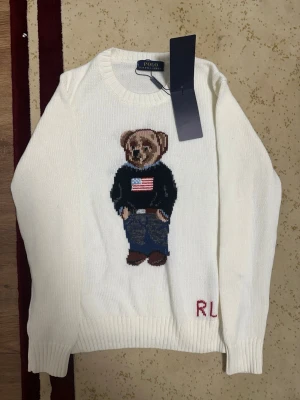 Vit stickad tröja Polo Ralph Lauren - Vit stickad tröja från Polo Ralph Lauren med ikonisk björn på bröstet, klädd i jeans och mörk tröja med amerikansk flagga. Rund halsringning och ribbade muddar. RL-broderi nertill. Perfekt för dig som gillar klassisk och lekfull stil.