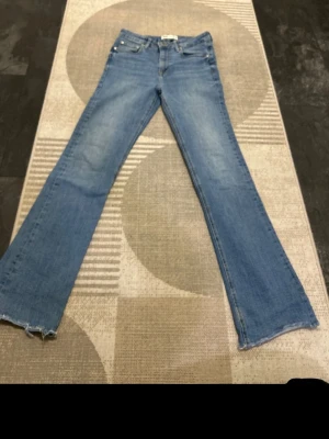 Blå bootcut jeans från Zara - Säljer ett par ljusblå bootcut jeans från Zara med klassisk femficksdesign och råa kanter vid bensluten. Jeansen har en snygg tvättad look och är tillverkade i mjukt denimtyg med lite stretch för extra komfort.