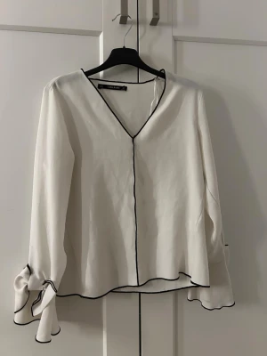 Vit blus med svarta detaljer från Zara - Stilren vit blus från Zara med svarta kontrastsömmar och v-ringning. Blusen har långa ärmar med dekorativa rosetter vid ärmsluten och lätt utsvängd passform. Perfekt för en trendig och chic look. Materialet känns mjukt och följsamt.