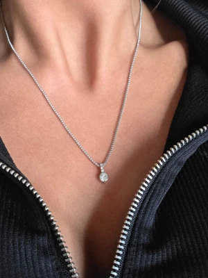 Silverfärgat halsband med glittrande sten - Snyggt halsband i silverfärg med en glittrande rund klar sten som hänge. Kedjan är boxlänk och hänget har små gnistrande stenar ovanför den stora stenen. Perfekt accessoar för att lyfta din outfit.