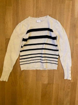 Vit stickad tröja med svarta ränder - Vit stickad tröja från Vila med svarta horisontella ränder över bröstet och magen. Tröjan har långa ärmar med puffdetalj vid axlarna och tre dekorativa knappar i guld och vitt vid ärmsluten. Ribbstickade muddar och rund hals.