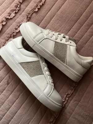 Vita sneakers med strassdetaljer OBS har en blå fläck se bilder !!! - Snygga vita sneakers med glittriga strassdetaljer på sidan. Skorna har klassisk låg modell, vita snören och en tjock sula. Tillverkade i syntetmaterial med en stilren och modern look som passar till många outfits. OBS har en blå fläck se bilder 