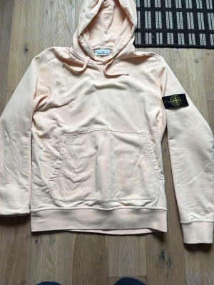 Ljusbeige hoodie från Stone Island - Säljer en ljusbeige hoodie från Stone Island med klassisk patch på vänster ärm. Hoodien har stor känguruficka framtill, ribbade muddar och en skön huva. Tillverkad i mjuk bomull som känns riktigt bekväm. Perfekt för alla stilar 