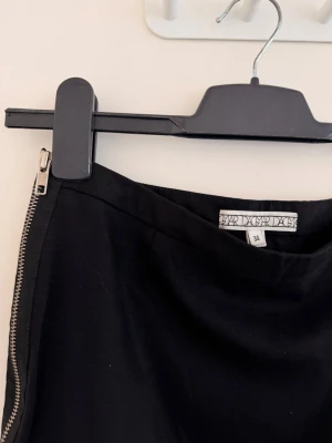 Dagmar Ull kjol med dragkedja- Storlek 34 - Fantastisk svart kjol från det svenska premiummärket House of Dagmar. Ett riktigt basplagg i garderoben med en kaxig twist, med en dekorativ silverfärgad YKK-dragkedja längs sidan. Notera: Dragkedjan är stängd nertill (inte ett slits), vilket fungerar som en snygg detalj och ger en högkvalitativ finish. Tillverkad av en förstklassig ullblandning (98 % ull) med en gnutta stretch för en perfekt, skräddarsydd passform. Minimalistisk skandinavisk design när den är som bäst.