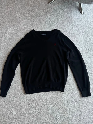 Svart stickad tröja från Ralph Lauren - Klassisk svart stickad tröja från Ralph Lauren med V-ringning och lång ärm. Tröjan har en liten röd broderad logga på bröstet och ribbade muddar vid ärmslut och nederkant. Perfekt att matcha med jeans eller kjol.