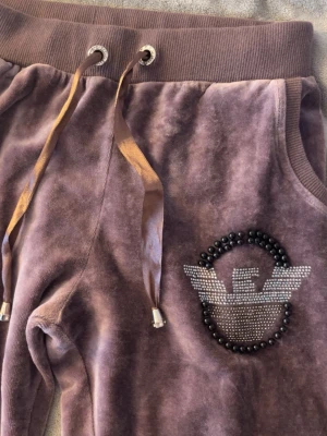 Lila mjukisbyxor från Emporio Armani - Säljer ett par lila mjukisbyxor från Emporio Armani i mjuk velour. Byxorna har resårmidja med breda snören och fickor. På ena benet finns ett stort glittrigt Armani-logotyp i strass och svarta pärlor. Perfekta för chill dagar eller när du vill vara extra comfy.