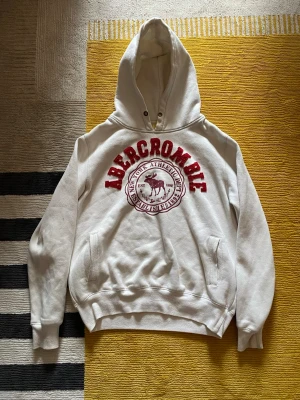 Vit hoodie från Abercrombie - Säljer en vit hoodie från Abercrombie med stor röd logga och tryck framtill. Hoodien har huva med dragsko, känguruficka och ribbade muddar vid ärmar och nederkant. Perfekt för dig som gillar klassisk college-stil och vill ha något bekvämt och snyggt.