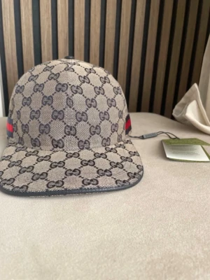 Gucci keps med GG-mönster  - Snygg keps från Gucci i beige canvas med klassiskt GG-monogram över hela kepsen. På sidan finns en ikonisk röd och blå rand. Kepsen har böjd skärm och justerbar passform. Perfekt för dig som vill ha en exklusiv accessoar. PRIS KAN DISKUTERAS!!