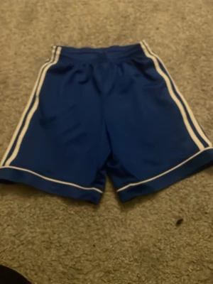 Blå Adidas shorts med vita ränder - Säljer ett par blå Adidas shorts med vita ränder längs sidorna och nederkanten. Shortsen har elastisk midja med snörning och är tillverkade i ett lätt och svalt material, perfekt för sport och träning.