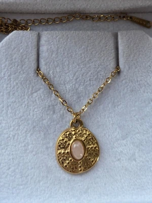 Guldigt halsband i rostfritt stål med rund berlock - Snyggt halsband i guldfärgat rostfritt stål med en rund, hamrad berlock. Berlocken har en ljus pärlliknande detalj i mitten som ger en elegant touch. Kedjan är tunn och justerbar för perfekt passform. Perfekt accessoar för att lyfta din outfit.