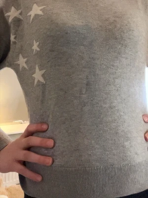 Grå stickad tröja med stjärnor och luva - Mysig grå stickad tröja med vita stjärnor på ena axeln och bröstet. Tröjan har rund halsringning, långa ärmar och ribbade muddar vid ärmslut och nederkant.