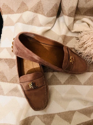 Beige mocka loafers från Ralph Lauren - Säljer ett par snygga bruna loafers i mocka från Ralph Lauren. Skorna har gummisula med nabbmönster och guldfärgad RL-logga på ovansidan. Klassisk design med rundad tå och sömmar runt kanten. Perfekta för en stilren look.