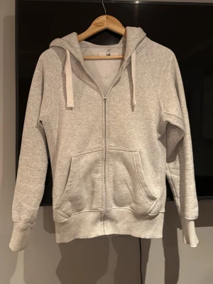 Ljusgrå zip-hoodie från Russemerch  - Ljusgrå hoodie med dragkedja från Russemerch storlek S. Mjuk fleece på insidan, stora fickor framtill och breda muddar vid ärmslut och nederkant. Huva med vita snören för justering. Perfekt för chill dagar och enkel att matcha med jeans eller joggers.