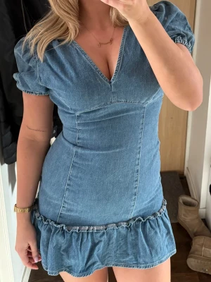 Jeansklänning med puffärm och volang - Trendig jeansklänning med kort puffärm, v-ringning och volangkant nedtill. Figurnära passform som framhäver siluetten. Perfekt för dig som gillar en avslappnad men ändå snygg look.