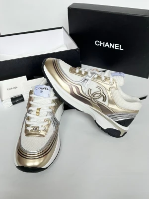 Chanel vita och guld sneakers - Snygga sneakers från Chanel i vitt och guld med metallicdetaljer och klassisk Chanel-logga på sidan. Ovandelen är i mesh och läder, med vita skosnören och robust sula i svart och vitt. Perfekta för dig som vill sticka ut med exklusiv stil.