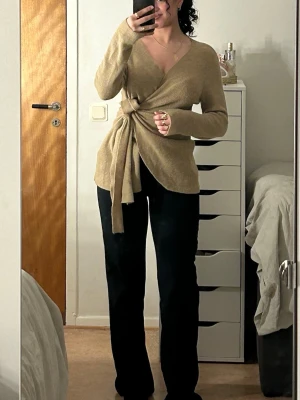 Beige omlottkofta med knytband - Trendig beige omlottkofta med långa ärmar och breda knytband i midjan. Koftan har en djup v-ringning och är gjord i ett mjukt, stickat material som ger en avslappnad men ändå stilren look. Perfekt att matcha med både jeans och kostymbyxor. Aldrig använt’