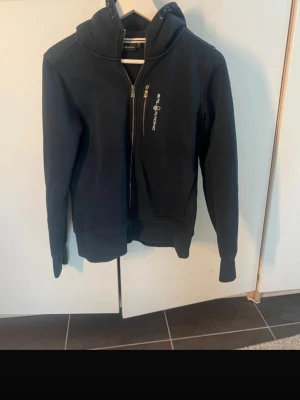 Mörkblå hoodie från Sail Racing - Snygg mörkblå hoodie från Sail Racing med dragkedja framtill och en bröstficka med logga. Hoodien har en stor huva och ribbade muddar vid ärmar och nederkant. Perfekt för dig som gillar stilrena och sportiga plagg.